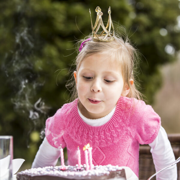 Ein junges Mädchen, das eine Krone trägt, bläst im Freien die Kerzen auf einer Geburtstagstorte aus, wobei Rauch von den erloschenen Kerzen aufsteigt.