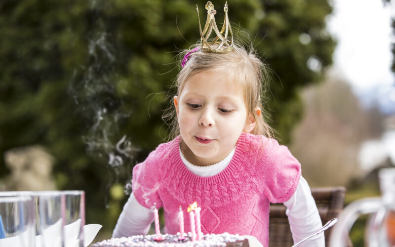 Ein junges Mädchen, das eine Krone trägt, bläst im Freien die Kerzen auf einer Geburtstagstorte aus, wobei Rauch von den erloschenen Kerzen aufsteigt.