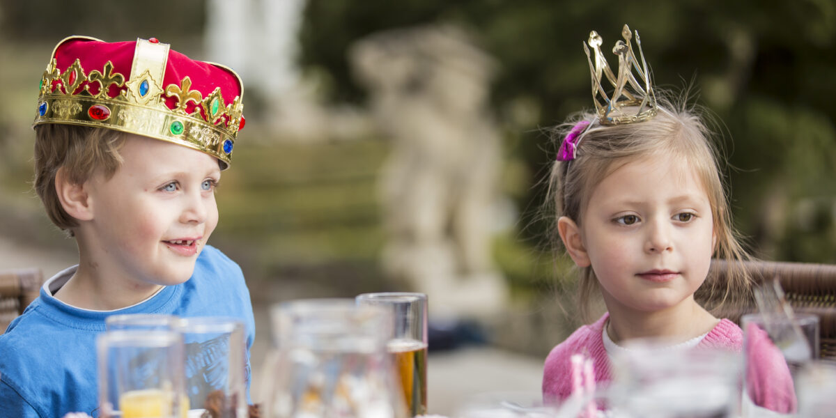 Zwei kleine Kinder mit Kronen sitzen an einem Tisch im Freien mit Getränken und Speisen, darunter Orangensaft und Kuchen, in einem Garten.