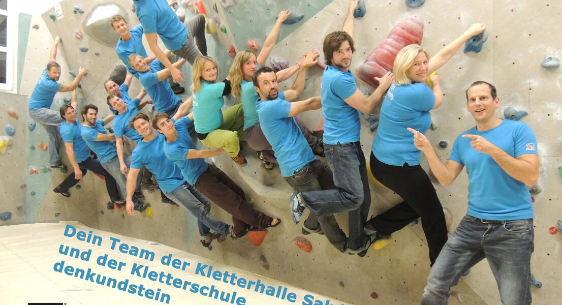 Eine Gruppe von Personen in passenden blauen Hemden posiert an einer Indoor-Kletterwand in der Kletterhalle Salzburg, mit Text und Logos für die Kletterhalle und Kletterschule am unteren Rand.