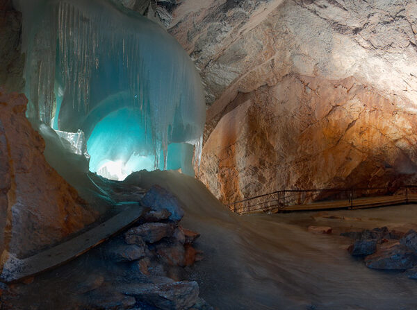 Große Eisformation in einer Felsenhöhle, die von blauem Licht beleuchtet wird, mit einem Weg und verstreuten Steinen auf dem Höhlenboden.