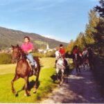 Vier Personen reiten auf einem unbefestigten Weg neben einem grasbewachsenen Feld, mit einem Dorf und bewaldeten Hügeln im Hintergrund unter einem klaren Himmel.