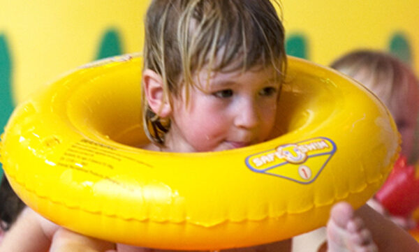 Ein kleines Kind mit nassen Haaren benutzt einen gelben aufblasbaren Schwimmring in einem Pool. Andere Kinder sind teilweise im Hintergrund zu sehen.