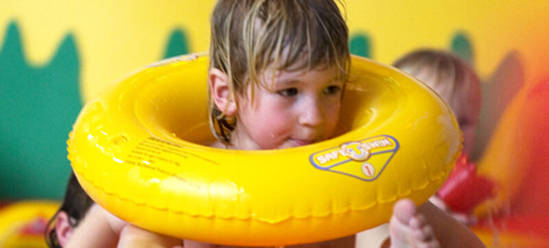 Ein kleines Kind mit nassen Haaren benutzt einen gelben aufblasbaren Schwimmring in einem Pool. Andere Kinder sind teilweise im Hintergrund zu sehen.