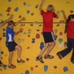 Vier Kinder klettern in Sportkleidung und Kletterschuhen seitwärts an einer gelben Indoor-Kletterwand mit verschiedenfarbigen Griffen.