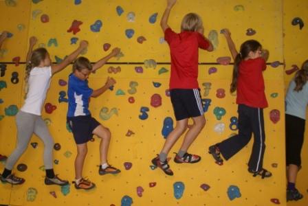 Vier Kinder klettern in Sportkleidung und Kletterschuhen seitwärts an einer gelben Indoor-Kletterwand mit verschiedenfarbigen Griffen.