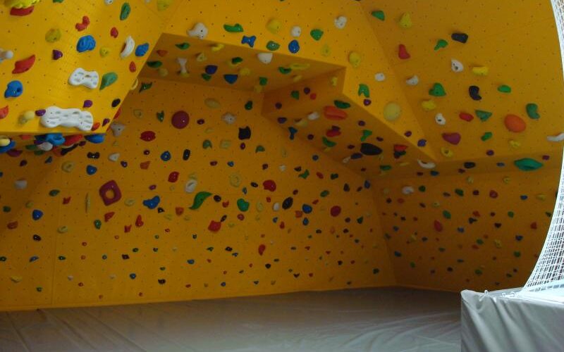 Indoor-Boulderwand mit gelben Platten und verschiedenfarbigen Klettergriffen, die mehrere Winkel abdecken, über einem gepolsterten Boden.