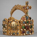 Eine mit Edelsteinen, Perlen und einem großen Kreuz geschmückte Goldkrone, die in der Mitte mit einem Bild und Inschriften verziert ist.