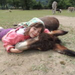 Ein junges Mädchen mit rosa Jacke und Kopftuch liegt im Gras und umarmt einen ruhenden Esel, während im Hintergrund mehrere andere Esel grasen.