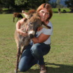Eine Frau kniet im Gras, lächelt und umarmt einen kleinen Esel im Freien mit Bäumen im Hintergrund.