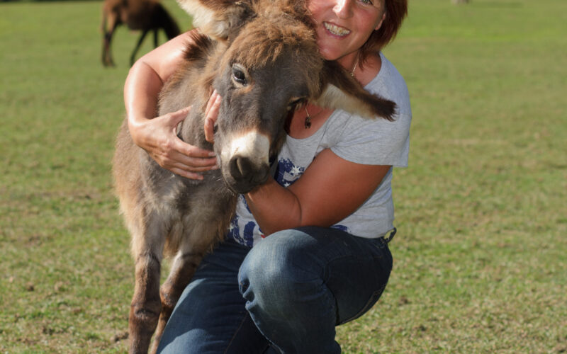 Eine Frau kniet im Gras, lächelt und umarmt einen kleinen Esel im Freien mit Bäumen im Hintergrund.