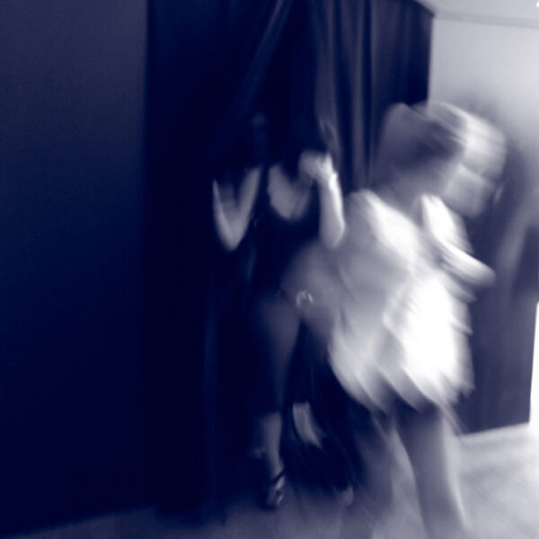 Ein unscharfes Foto zeigt drei Personen in Bewegung, von denen eine durch einen dunklen Vorhang austritt und zwei andere vorbeigehen; unten links erscheint der Schriftzug "S I N N E".