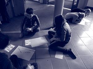 Vier Kinder sitzen in Gruppen auf dem Boden und arbeiten mit Stiften und Zetteln in einem gekachelten Innenraum an Papieren.