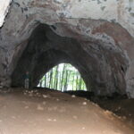 Eine Person steht in einer großen, felsigen Höhle mit einer weit gewölbten Öffnung, durch die Bäume und Tageslicht nach draußen fallen.