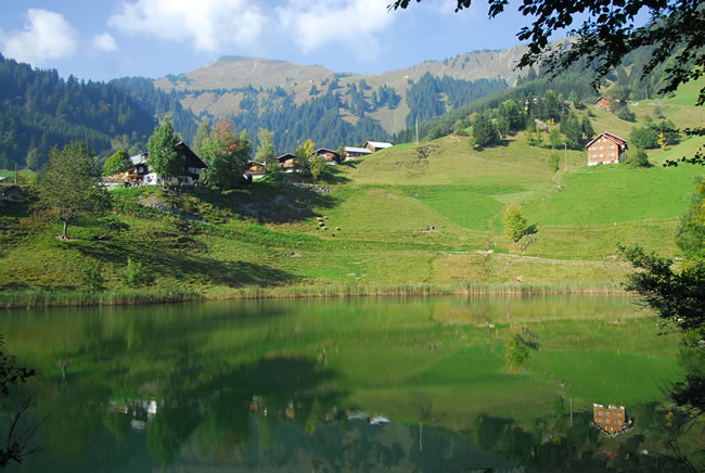 In einem ruhigen See spiegeln sich grüne Hügel, verstreute Häuser und bewaldete Berge unter einem teilweise bewölkten Himmel.