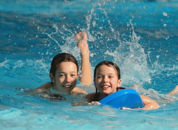 Zwei Kinder schwimmen in einem Pool und lächeln in die Kamera, wobei ein Kind ein blaues Kickboard hält und das Wasser hinter ihnen spritzt.