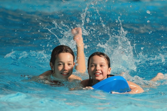 Zwei Kinder schwimmen in einem Pool und lächeln in die Kamera, wobei ein Kind ein blaues Kickboard hält und das Wasser hinter ihnen spritzt.