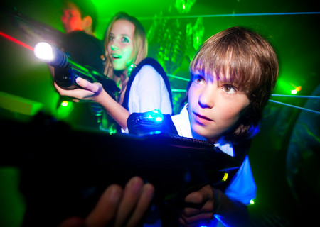 Zwei Kinder spielen in einem dunklen Raum mit Neonlicht Laser-Tag, zielen mit ihren Laserpistolen nach vorne und tragen Sensorwesten.