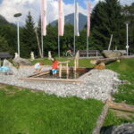 Zwei Kinder spielen in einem flachen, felsigen Wasserspiel in einem Park mit Bänken, Fahnen und einer Kulisse aus Bäumen und Bergen.