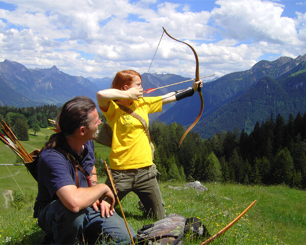 Ein Kind spannt einen Bogen und zielt einen Pfeil, während ein Erwachsener zuschaut. Beide befinden sich im Freien auf einem grasbewachsenen Hügel mit Bergen und Bäumen im Hintergrund unter einem teilweise bewölkten Himmel.