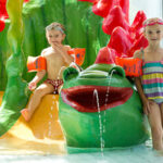 Zwei kleine Kinder in Badeanzügen und Schwimmflügeln stehen und sitzen neben einem Wasserspiel mit Froschmotiv in einem Hallenbad.