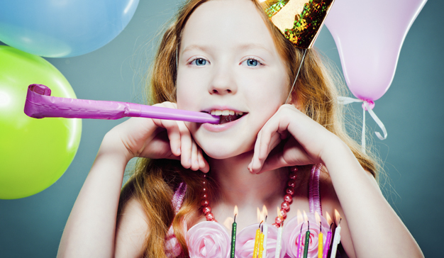 Ein junges Mädchen mit langen roten Haaren trägt einen Partyhut und hält eine Partytorte im Mund. Vor ihr stehen Luftballons und eine Geburtstagstorte mit brennenden Kerzen.