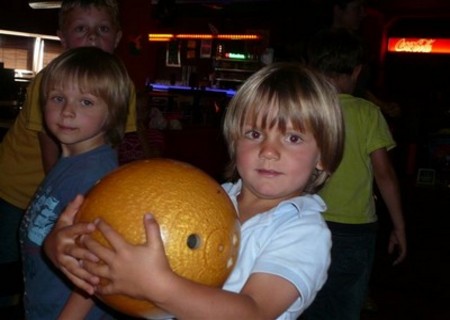 Im Vordergrund steht ein kleines Kind, das eine gelbe Bowlingkugel in der Hand hält, im Hintergrund sind andere Kinder und eine schwach beleuchtete Bowlingbahn zu sehen.