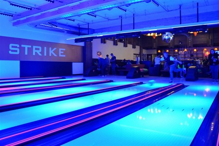 Neonbeleuchtete Bowlingbahnen mit einem großen "STRIKE"-Schild an der Wand; im Lounge-Bereich im Hintergrund sind Menschen versammelt und unterhalten sich.