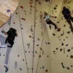 Drei Personen mit Klettergurten klettern an einer Indoor-Kletterwand mit farbigen Griffen und Sicherungsseilen.