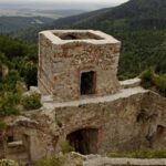 Ruinen eines alten Steingebäudes mit einem quadratischen Turm, umgeben von Grün und bewaldeten Hügeln im Hintergrund.