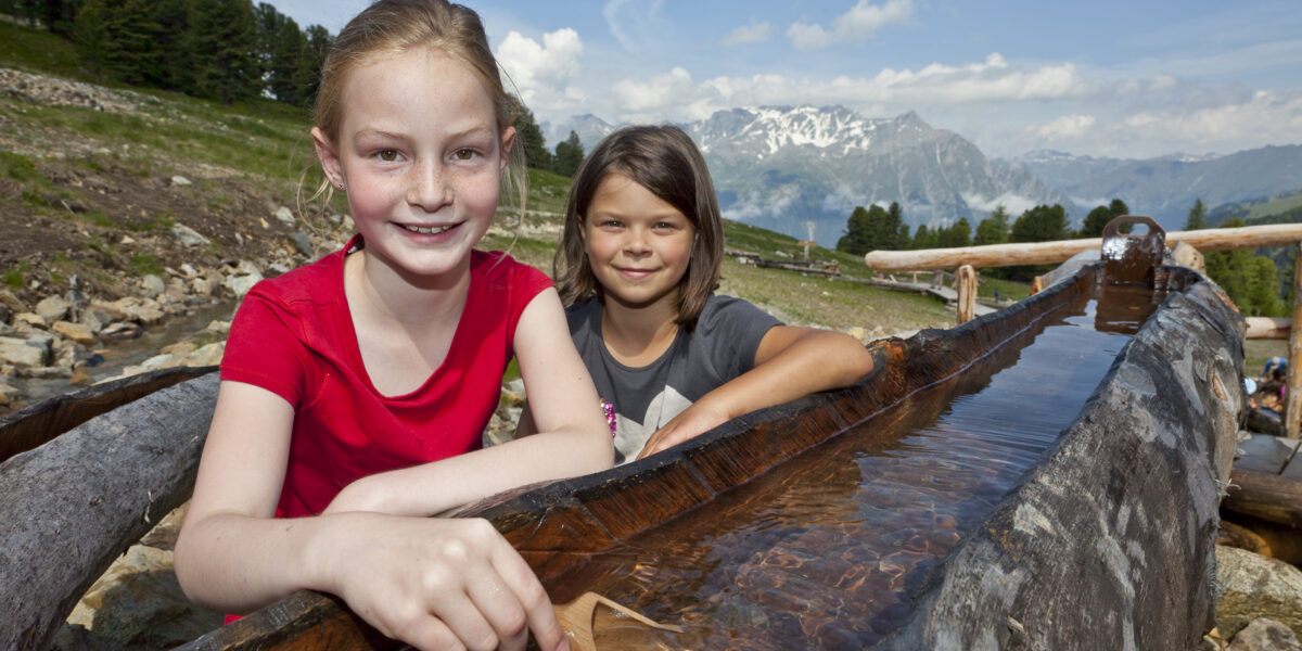 Zwei Mädchen spielen an einem hölzernen Wasserkanal im Freien, mit Bergen und Bäumen im Hintergrund unter einem teilweise bewölkten Himmel.