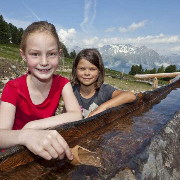 Zwei Mädchen spielen an einem hölzernen Wasserkanal im Freien, mit Bergen und Bäumen im Hintergrund unter einem teilweise bewölkten Himmel.