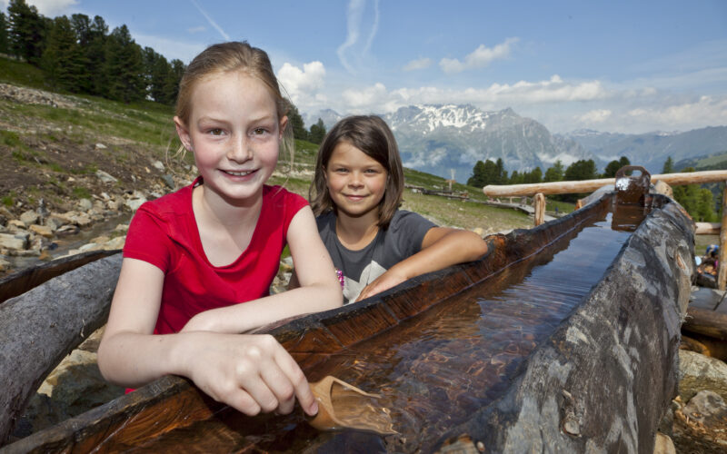 Zwei Mädchen spielen an einem hölzernen Wasserkanal im Freien, mit Bergen und Bäumen im Hintergrund unter einem teilweise bewölkten Himmel.