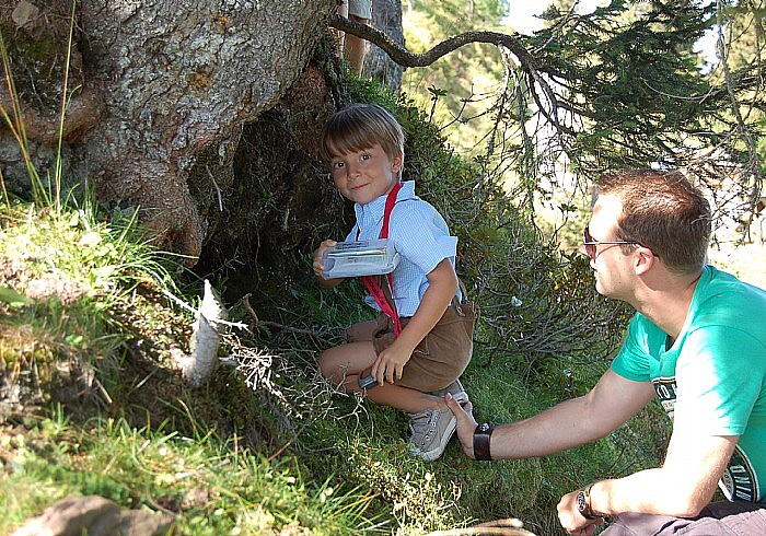 Ein kleiner Junge kauert an einem Baum und hält einen Behälter in der Hand, während ein erwachsener Mann mit Sonnenbrille neben ihm auf einer Wiese kniet.