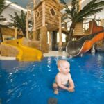 Das Baby sitzt und planscht im flachen Innenpoolbereich mit Rutschen, Spielgeräten und künstlichen Palmen im Hintergrund.