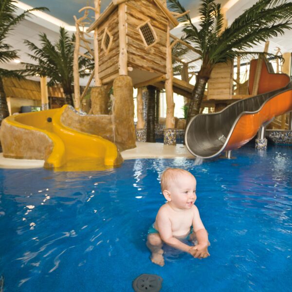 Das Baby sitzt und planscht im flachen Innenpoolbereich mit Rutschen, Spielgeräten und künstlichen Palmen im Hintergrund.