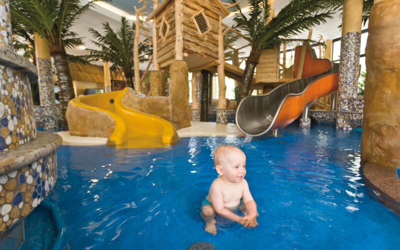 Das Baby sitzt und planscht im flachen Innenpoolbereich mit Rutschen, Spielgeräten und künstlichen Palmen im Hintergrund.