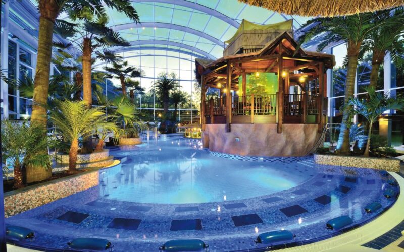 Tropischer Innenpool mit Palmen, blauen Kacheln und einem zentralen Holzpavillon unter einer großen Glaskuppel.