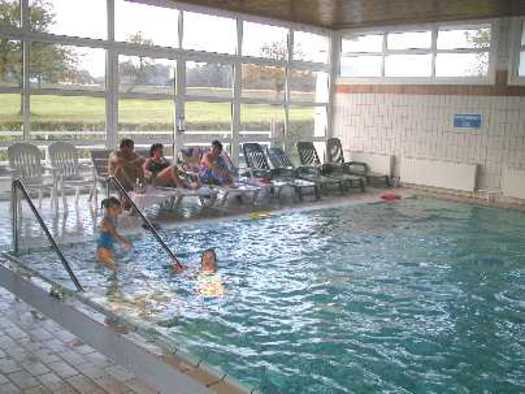 Mehrere Personen sitzen auf Liegestühlen und schwimmen in einem Hallenbad mit großen Fenstern und gefliestem Boden.