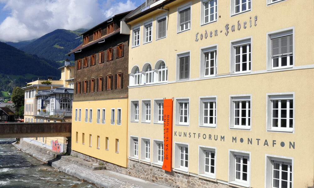 Ein gelbes Gebäude mit der Aufschrift "Kunstforum Montafon" steht neben einem fließenden Fluss, mit Bergen und einem Steg im Hintergrund unter einem teilweise bewölkten Himmel.