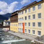 Ein gelbes Gebäude mit der Aufschrift "Kunstforum Montafon" steht neben einem fließenden Fluss, mit Bergen und einem Steg im Hintergrund unter einem teilweise bewölkten Himmel.