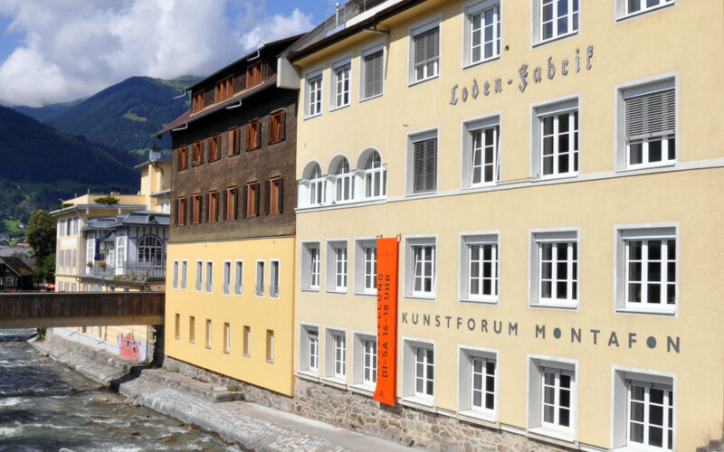 Ein gelbes Gebäude mit der Aufschrift "Kunstforum Montafon" steht neben einem fließenden Fluss, mit Bergen und einem Steg im Hintergrund unter einem teilweise bewölkten Himmel.