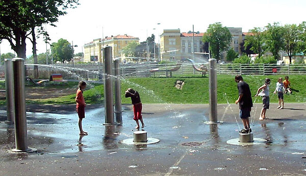 Kinder spielen an einem sonnigen Tag in einem Park in einem Planschbecken im Freien und stehen auf Plattformen, während Wasser von Metallsäulen spritzt.