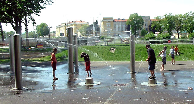 Kinder spielen an einem sonnigen Tag in einem Park in einem Planschbecken im Freien und stehen auf Plattformen, während Wasser von Metallsäulen spritzt.