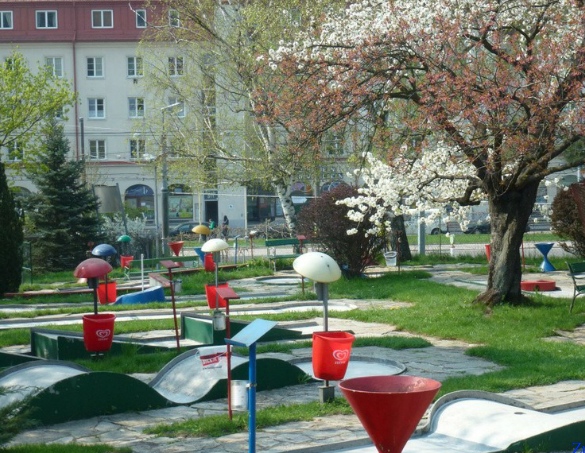 Ein Minigolfplatz mit bunten Hindernissen und roten Mülleimern, umgeben von Gras, Bäumen und blühenden Blumen, mit Gebäuden im Hintergrund.