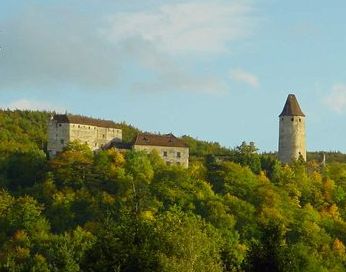 Ein steinernes Schloss mit einem angrenzenden hohen Turm steht auf einem baumbestandenen Hügel unter einem teilweise bewölkten Himmel.