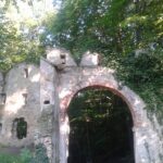 Ein teilweise verfallener steinerner Torbogen und eine mit Efeu überwucherte Mauer stehen in einem Waldgebiet, in dem das Sonnenlicht durch die Bäume dringt.