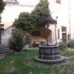 Ein steinerner Brunnen mit einer dekorativen Metallabdeckung steht in einem grasbewachsenen Innenhof neben einer Treppe, umgeben von alten Stuckmauern und Grünpflanzen.