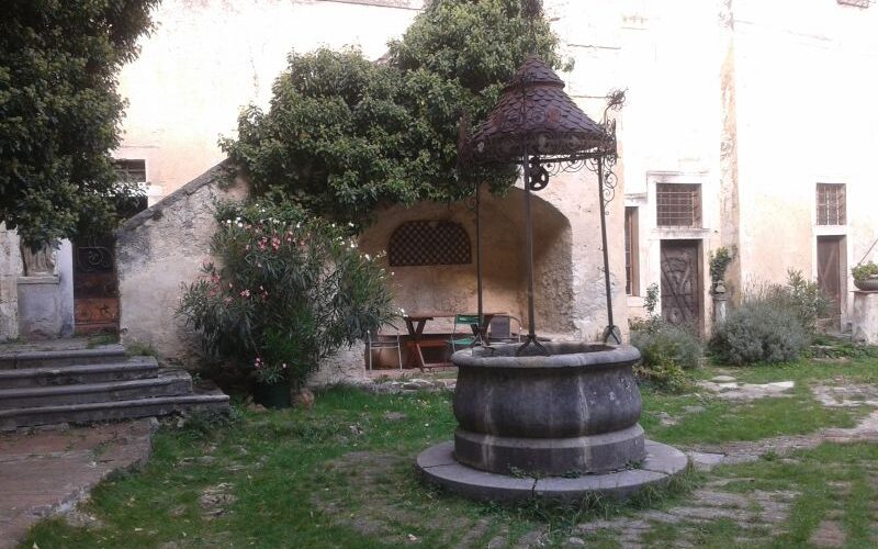 Ein steinerner Brunnen mit einer dekorativen Metallabdeckung steht in einem grasbewachsenen Innenhof neben einer Treppe, umgeben von alten Stuckmauern und Grünpflanzen.