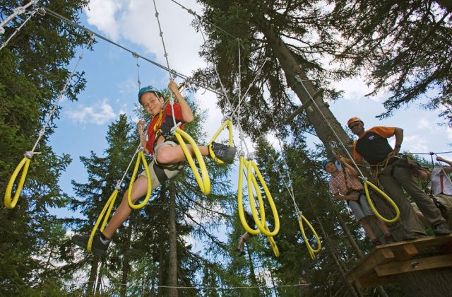 Ein Kind, das einen Helm und einen Klettergurt trägt, durchquert einen Seilparcours in einem Wald, während drei Personen von einer Holzplattform aus zusehen.